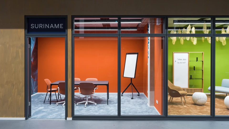 View through the glass front into the colorful meeting room 'Suriname' featuring an orange wall and a flipchart.