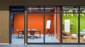 View through the glass front into the colorful meeting room 'Suriname' featuring an orange wall and a flipchart.