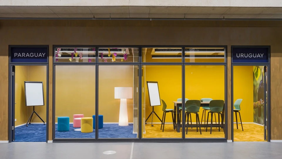 Meeting room in smartvillage Cologne-Muelheim featuring yellow walls, light blue chairs, and a light wooden table.