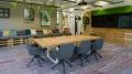 A modern meeting room featuring a large wooden table surrounded by six grey chairs, decorated with green plants and artwork on the walls.