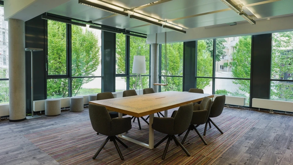 Modern meeting room featuring a large wooden table surrounded by dark chairs, expansive windows, and a view of greenery outside.