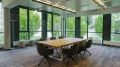 Modern meeting room featuring a large wooden table surrounded by dark chairs, expansive windows, and a view of greenery outside.