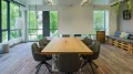 Modern meeting room with a large wooden table, surrounded by gray chairs, white screen on the wall, large windows, and green plants