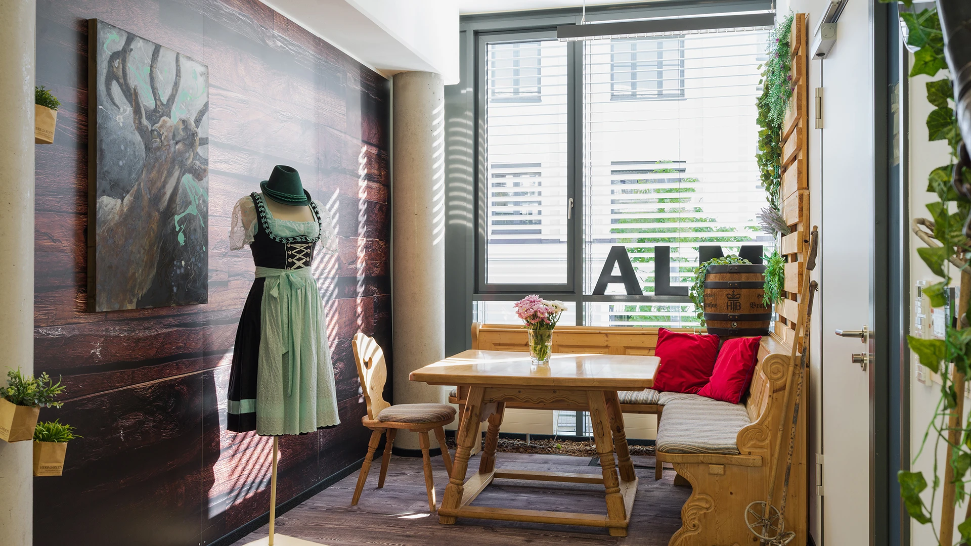 Cozy, bright room featuring a large wall painting of a deer, a rustic wooden table, chairs, a mannequin dressed in a green dress and hat, and numerous plants.