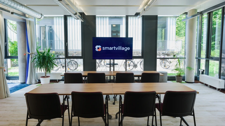 Conference room with a long wooden table and red chairs, a screen with the 'smartvillage' logo at the front, large windows in the background.