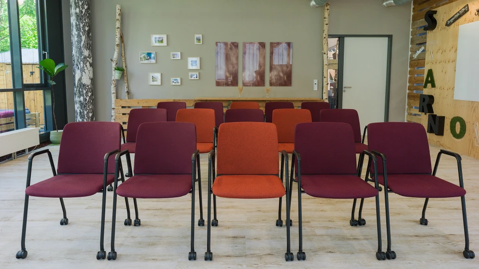 A seminar room with several rows of red and orange chairs facing a whiteboard, surrounded by plants and decorations on the walls.