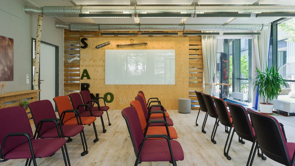Modern seminar room interior with wooden walls, red and black chairs, a projector screen, and plants near the windows.
