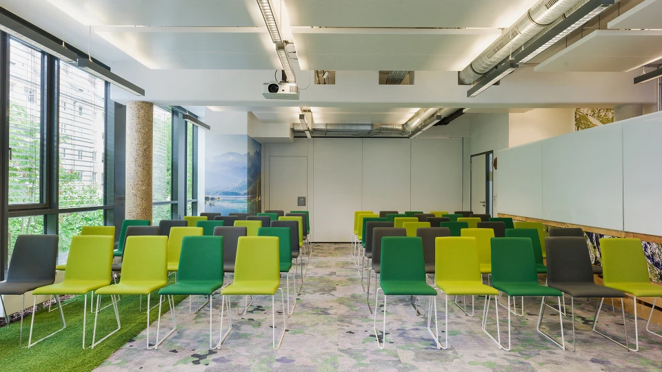 Modernes Seminarraum-Interieur mit Reihen grüner und grauer Stühle, Teppichboden mit buntem Muster und großen Fenstern mit Blick auf eine Berglandschaft.