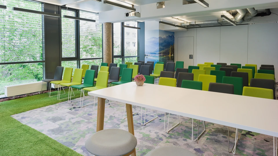 Modernes Bürokonferenzzimmer mit grünen und gelben Stühlen, einem großen weißen Tisch, einem Teppich mit Blumenmuster und großen Fenstern mit Blick auf grüne Pflanzen.