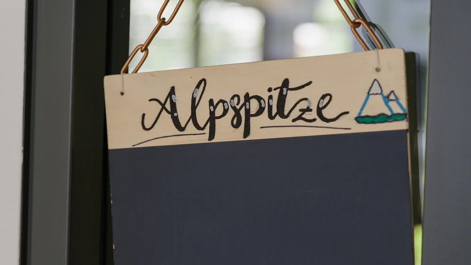 Holzschild mit der Aufschrift 'Alpspitze' und einer stilisierten Bergzeichnung, aufgehängt an einer Tür mit Glasfenster.