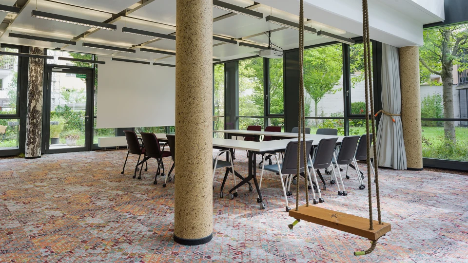 Modern office with a long conference table surrounded by chairs, a swing, and large windows overlooking green trees.