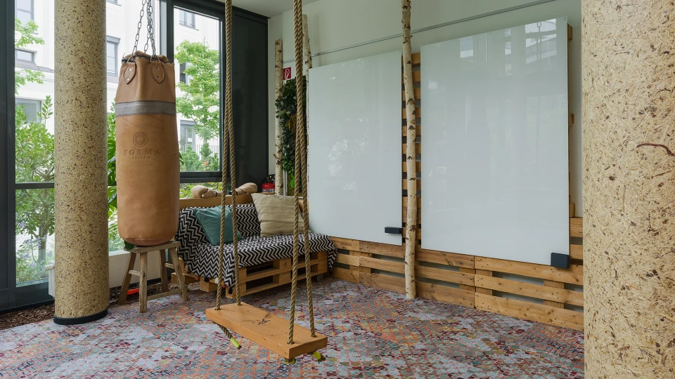 A bright room with large windows featuring a wooden swing, a punching bag, and a padded bench. The floor is covered with a colorful carpet.