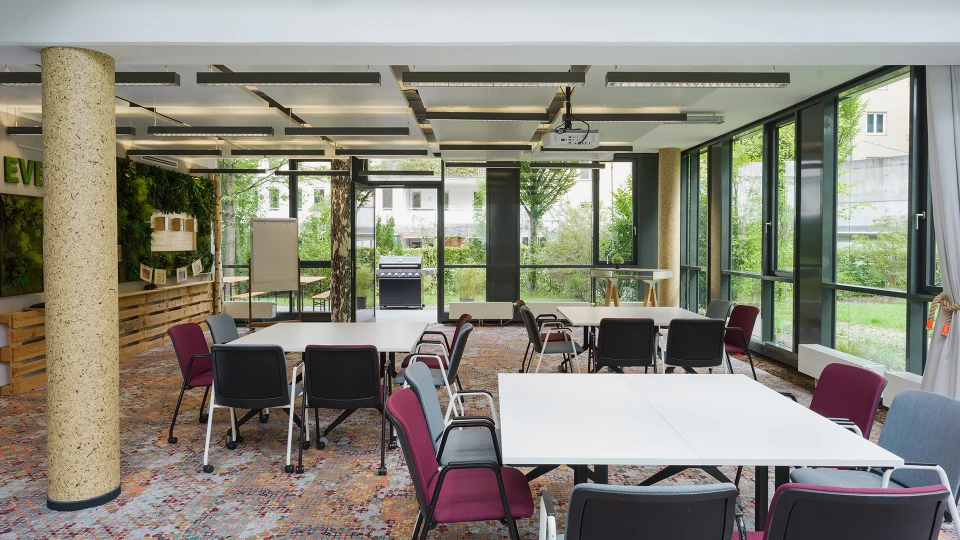 Bright seminar room with large windows, multiple tables and chairs. A whiteboard and a green plant wall are visible.