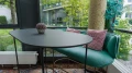 A modern rest area featuring an oval gray table, green velvet couch, and red pillows, positioned in front of large windows overlooking a green outdoor area.