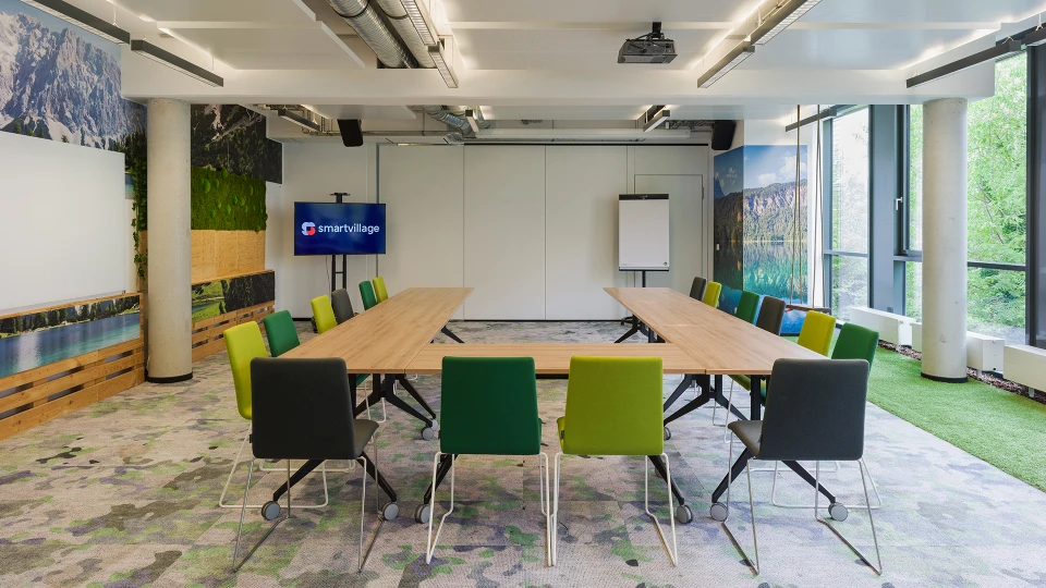 Modernes Besprechungszimmer mit einem langen Holztisch, umgeben von grünen und blauen Stühlen, großflächigen Bildern von Landschaften an den Wänden und einem Projektorbildschirm