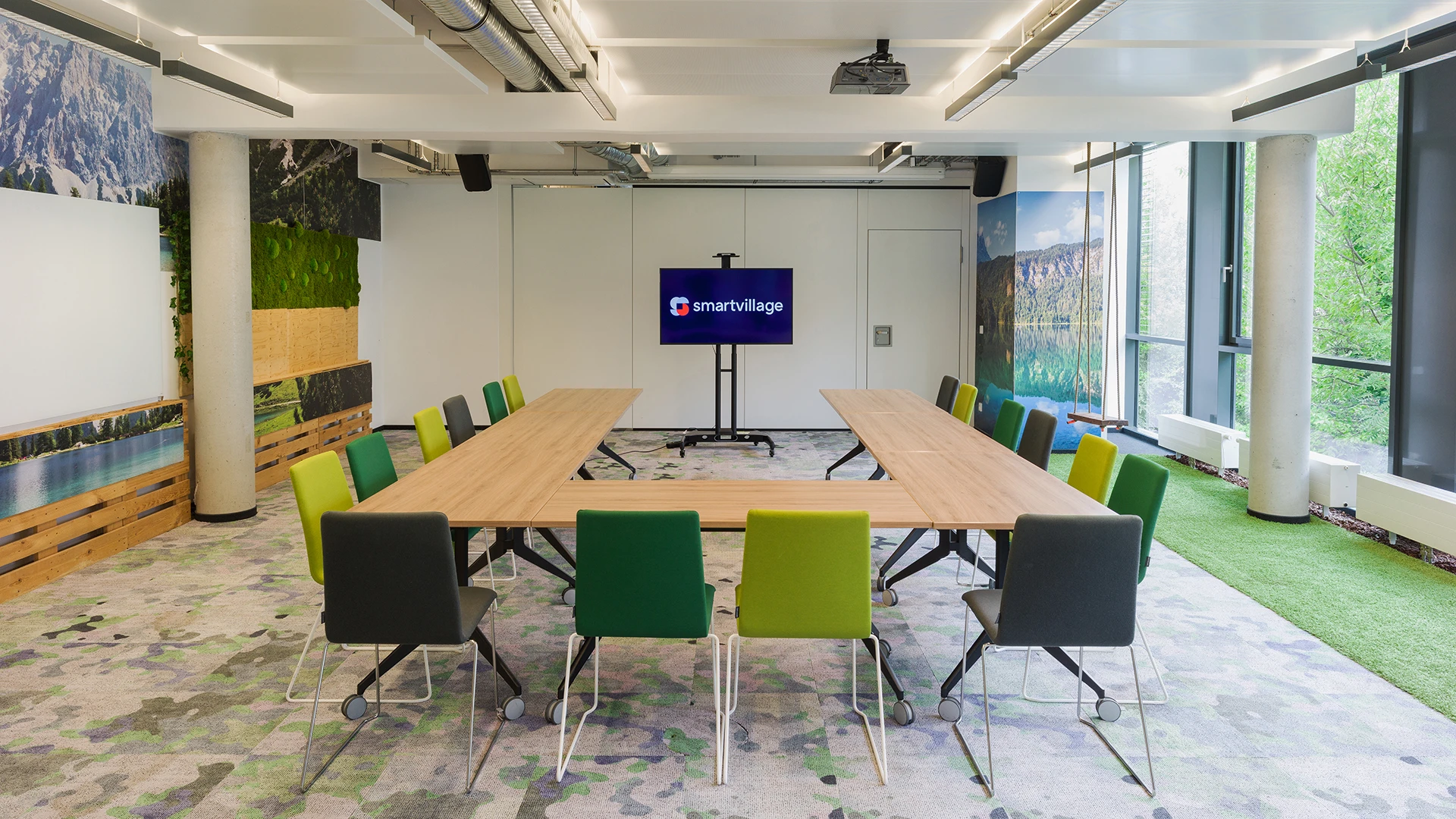Moderne Konferenzraumansicht mit einem großen Holztisch, umgeben von grünen und grauen Stühlen, einer Wand mit Berglandschaftsbild und einem Bildschirm mit dem Text 'teamwork'