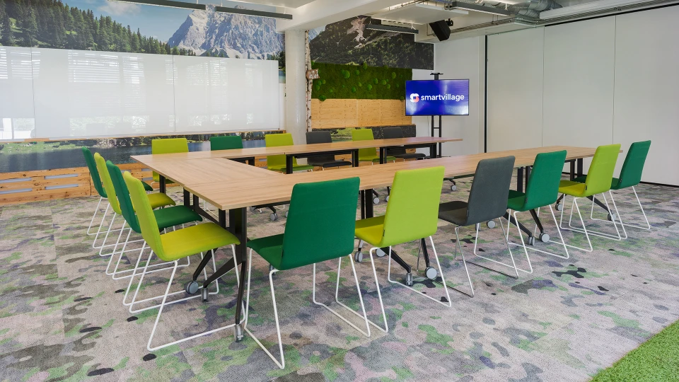 Moderner Besprechungsraum mit einem großen Holztisch, umgeben von grünen und grauen Stühlen, auf einem Teppich mit Naturmuster. Im Hintergrund eine Wand mit Bergpanorama und ein Bildschirm mit dem Logo 'smartenlight'.