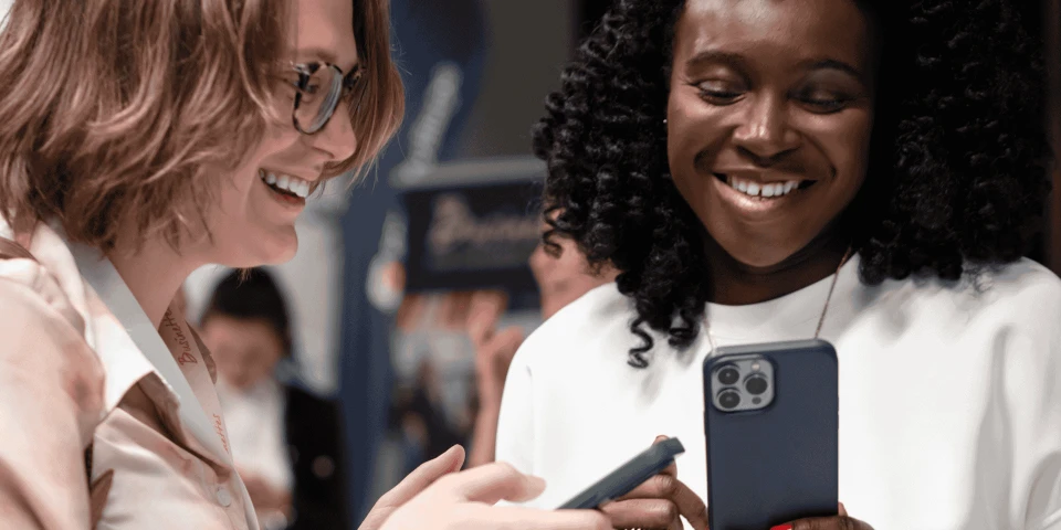 Two smiling women, one with curly hair and the other wearing glasses, looking at a smartphone in a busy conference setting
