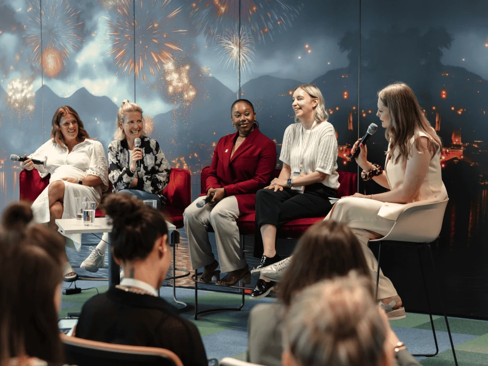 Five women sitting on stage discussing in front of an audience, with a backdrop of fireworks and city skyline.
