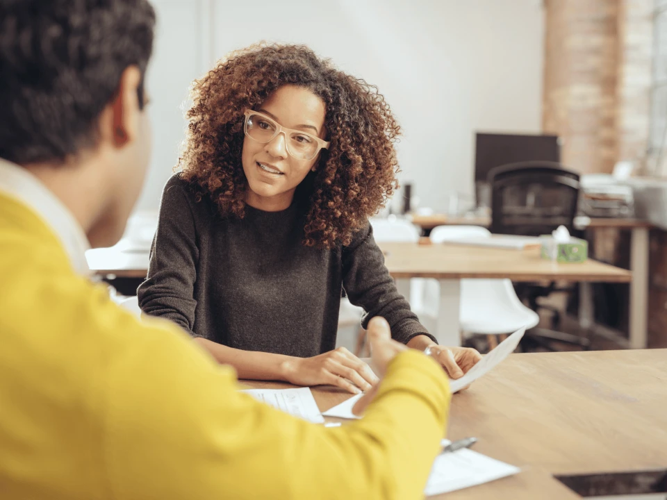 A woman with curly hair and glasses sitting at a wooden table having a discussion with a person whose back is visible.