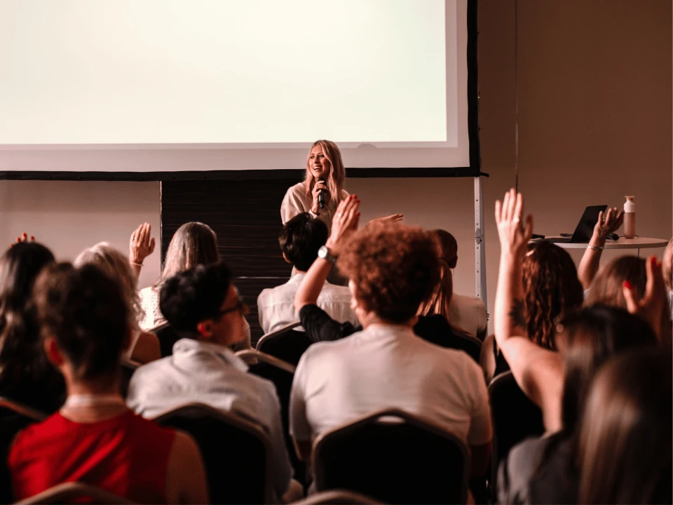 Woman standing in front of an audience in a seminar room speaking, with several people in the audience raising their hands