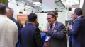 Mehrere Geschäftsleute unterhalten sich aktiv auf einer Messe mit blauem Teppich und Informationsständen im Hintergrund.