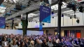 Publikum sitzt in einer modernen Halle mit Glasdach bei einer Konferenz, umgeben von Bannern und technischer Beleuchtung.
