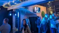 Besucher bei einem abendlichen Event in einer Bar mit Fernsehbildschirm, Beleuchtung und Gruppen von Menschen, die sich unterhalten.