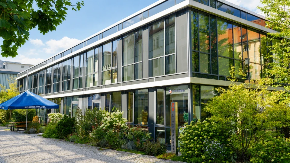 Modernes zweistöckiges Glasgebäude umgeben von grünen Bäumen und Sträuchern mit einem blauen Sonnenschirm im Vordergrund.