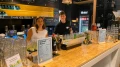 Zwei junge Mitarbeiter stehen hinter einer gut ausgestatteten Bar mit Gläsern, Getränken und einer Cocktailkarte. Hintergrund zeigt eine beleuchtete Tafel mit Menüoptionen.