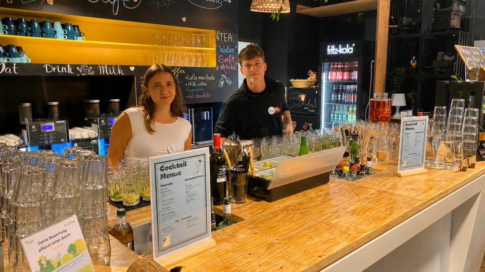 Zwei junge Mitarbeiter stehen hinter einer gut ausgestatteten Bar mit Gläsern, Getränken und einer Cocktailkarte. Hintergrund zeigt eine beleuchtete Tafel mit Menüoptionen.