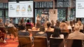 Eine Präsentatorin spricht vor einem Publikum von überwiegend weiblichen Zuhörern in einem modernen Konferenzraum mit großen Fenstern und Präsentationsbildschirmen, die 'Die drei Phasen' anzeigen.