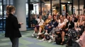 Junge Frau hält eine Präsentation vor einer sitzenden Gruppe von Zuhörerinnen in einem modernen Bürogebäude.