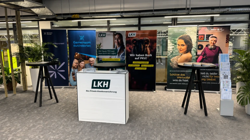 Ein Messestand mit mehreren Werbebannern, darunter eines für eine private Krankenversicherung, im Innenbereich mit einem Empfangstresen und dekorativen Pflanzen.