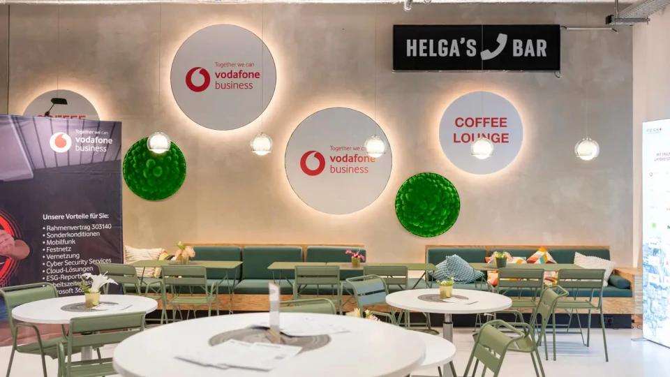 Innenansicht einer modernen Bar mit grünen Sitzgelegenheiten, weißen Tischen und Wänden mit Werbeplakaten von Vodafone. Über der Bar steht 'HELGA'S BAR'.
