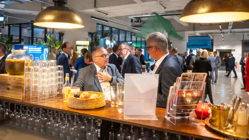 Zwei ältere Männer unterhalten sich an einer Bar bei einer Netzwerkveranstaltung. Im Hintergrund sind weitere Personen und Buffet-Elemente zu sehen.