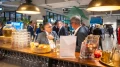 Zwei ältere Männer unterhalten sich an einer Bar bei einer Netzwerkveranstaltung. Im Hintergrund sind weitere Personen und Buffet-Elemente zu sehen.