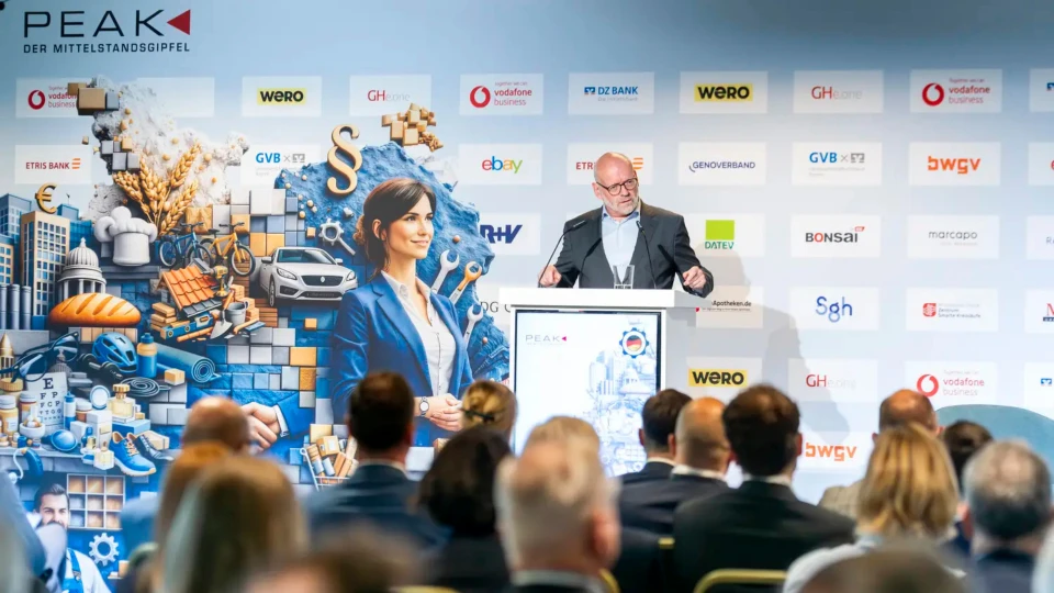 Ein Mann steht an einem Rednerpult auf einer Konferenz und spricht zu einem Publikum. Im Hintergrund sind verschiedene Unternehmenslogos und eine großformatige Grafik mit einer Frau zu sehen.