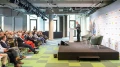 Ein Redner präsentiert auf einer Bühne vor einem Publikum in einem modernen Konferenzraum.