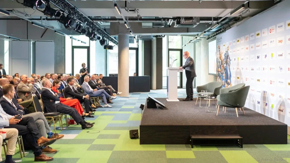 Ein Redner präsentiert auf einer Bühne vor einem Publikum in einem modernen Konferenzraum.