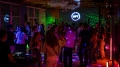 Menschen tanzen und unterhalten sich in einem lebhaften Nachtclub mit bunten Lichtern und einem DJ-Pult.