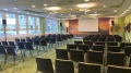 Leerer Konferenzraum mit Reihen von schwarzen Stühlen, die auf eine große Leinwand an der Vorderseite gerichtet sind. Der Raum hat grünen Teppich und Tageslicht durch Fenster.